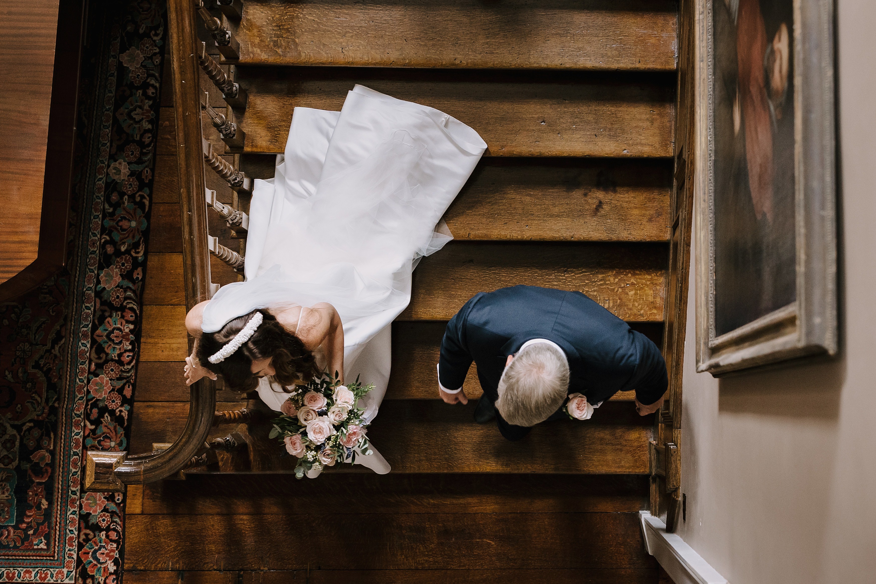 Photo from above showing a bride and father of the bride walking down the stairs together showing the train on her wedding dress
