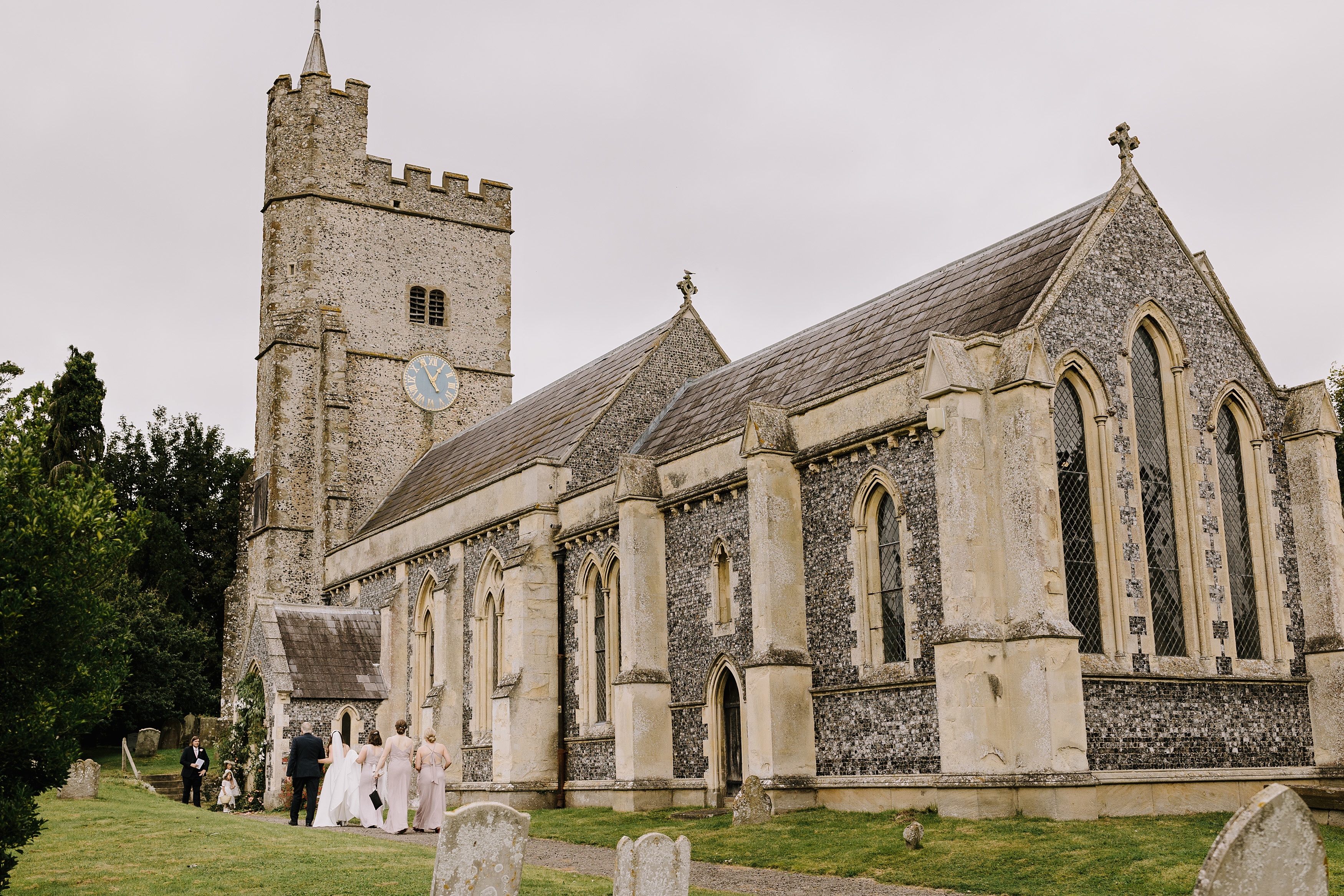 View of The Church of Holy Cross in Goodnestone with bride, bridesmaids, flower girls and father of the bride walking in