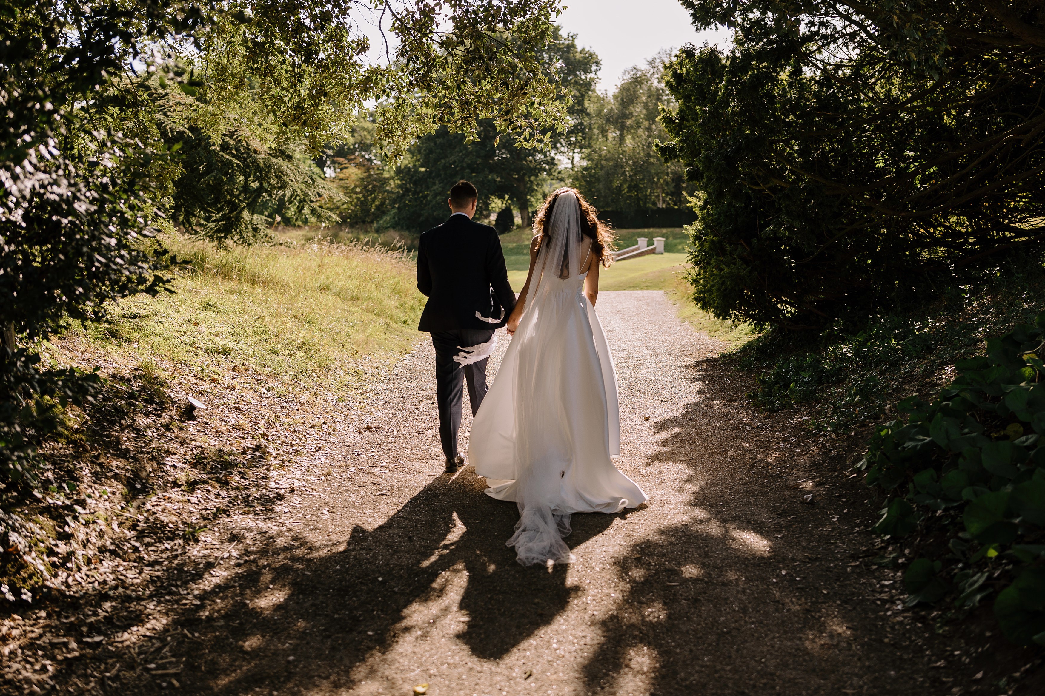 A photo from behind showing a bride and groom walk through Goodnestone Park hand in hand